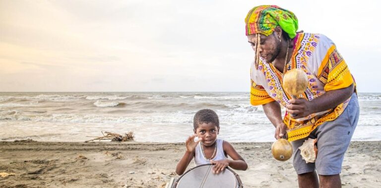 A child playing a drum beside a Garifuna musician holding maracas